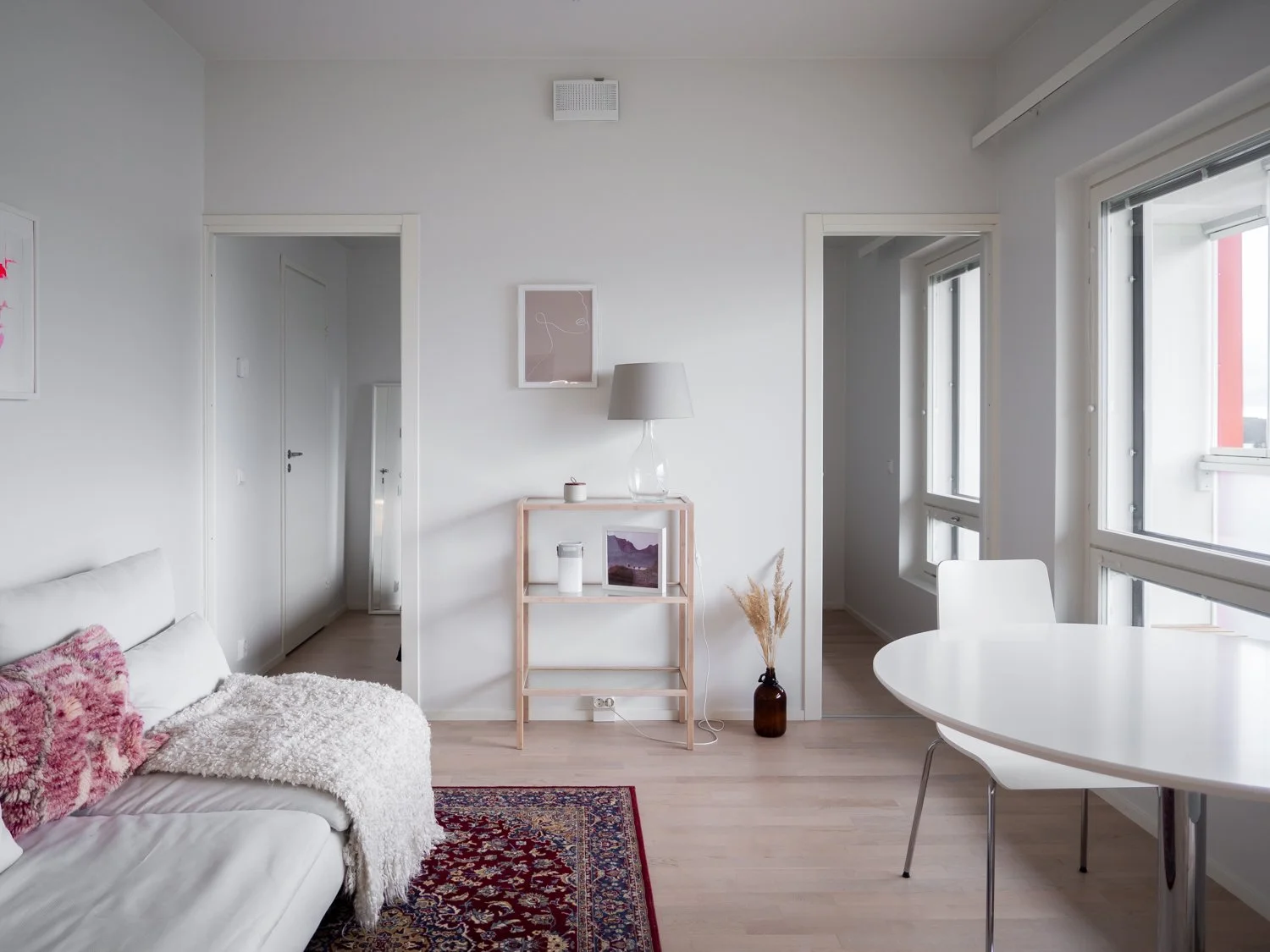 Minimalist living room with neutral tones, white sofa with pink cushions, decorative rug, wooden shelf with a lamp, small table, and large windows.