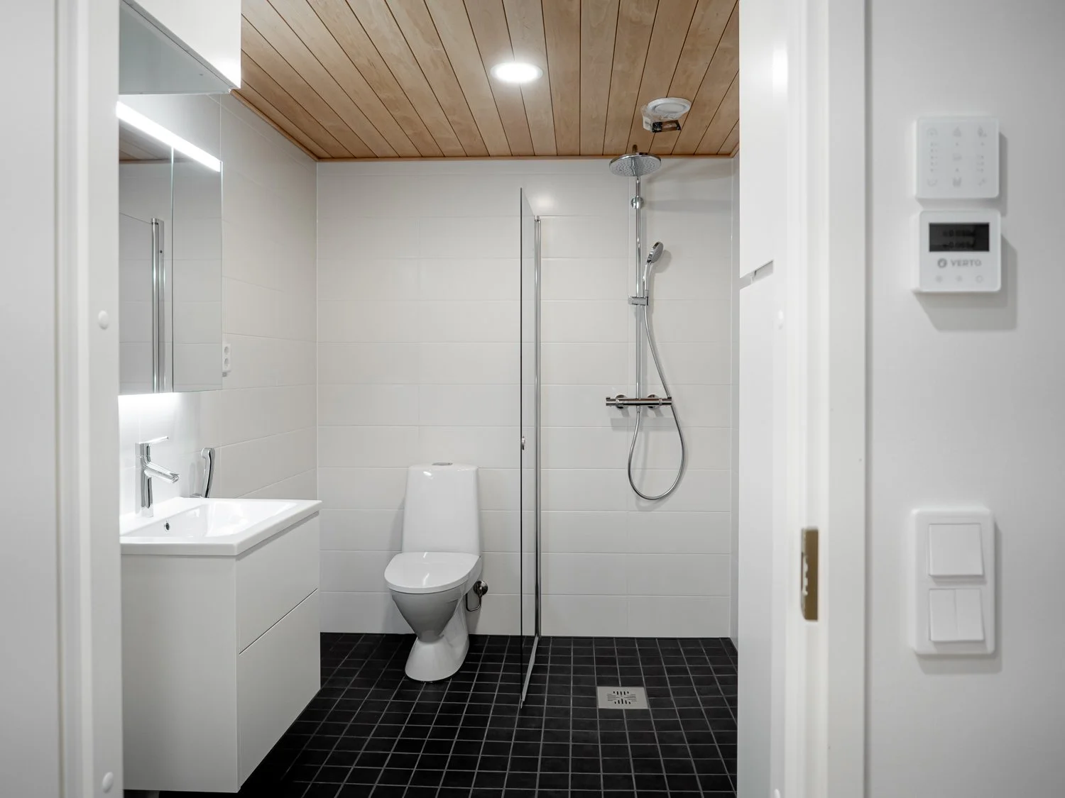 Modern bathroom with white tiles, wooden ceiling, wall-mounted toilet, sink with cabinet and mirror, and shower area.