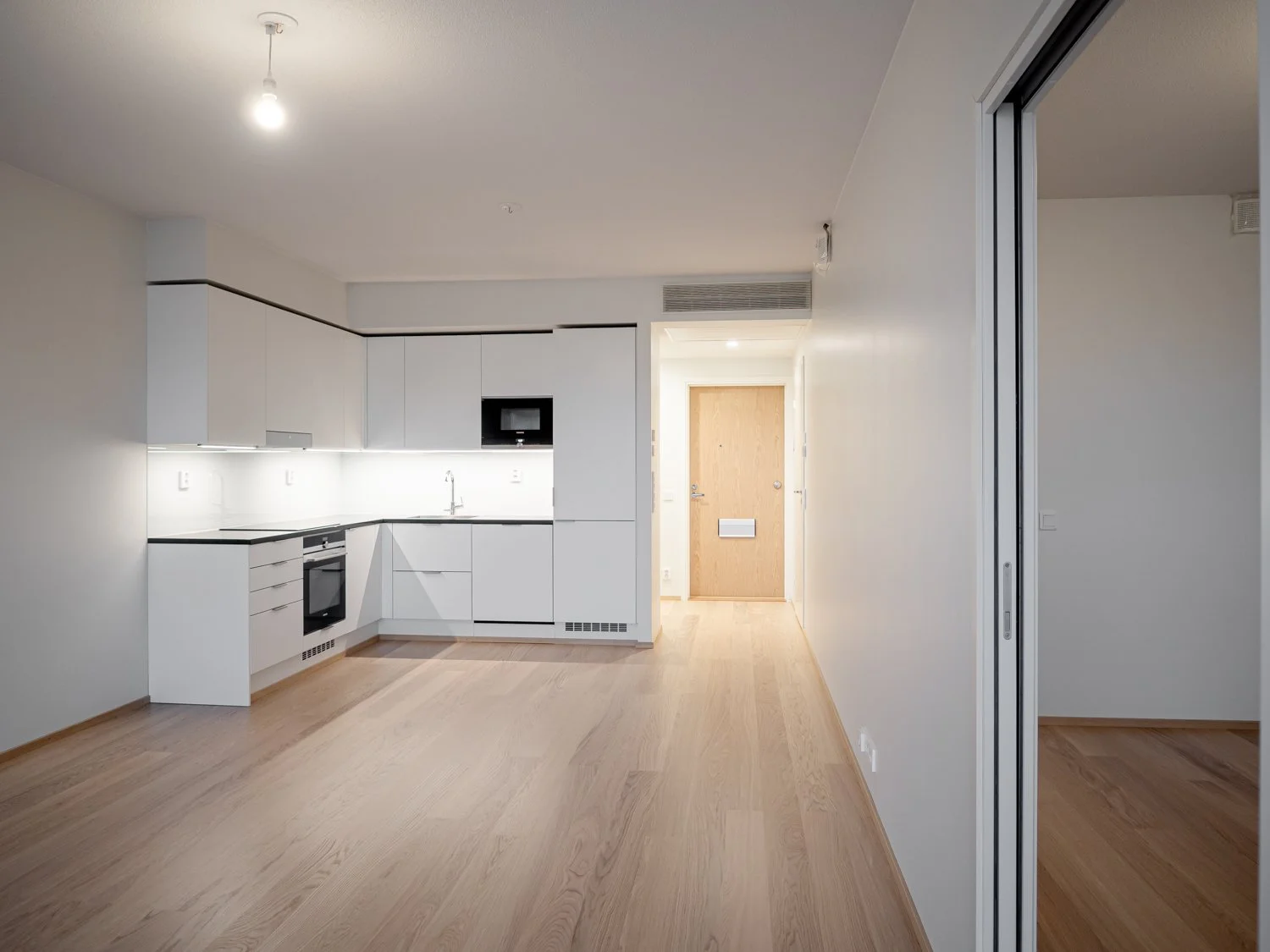 Modern minimalist kitchen with white cabinets, built-in appliances, and wooden flooring in an empty apartment space with bright lighting.
