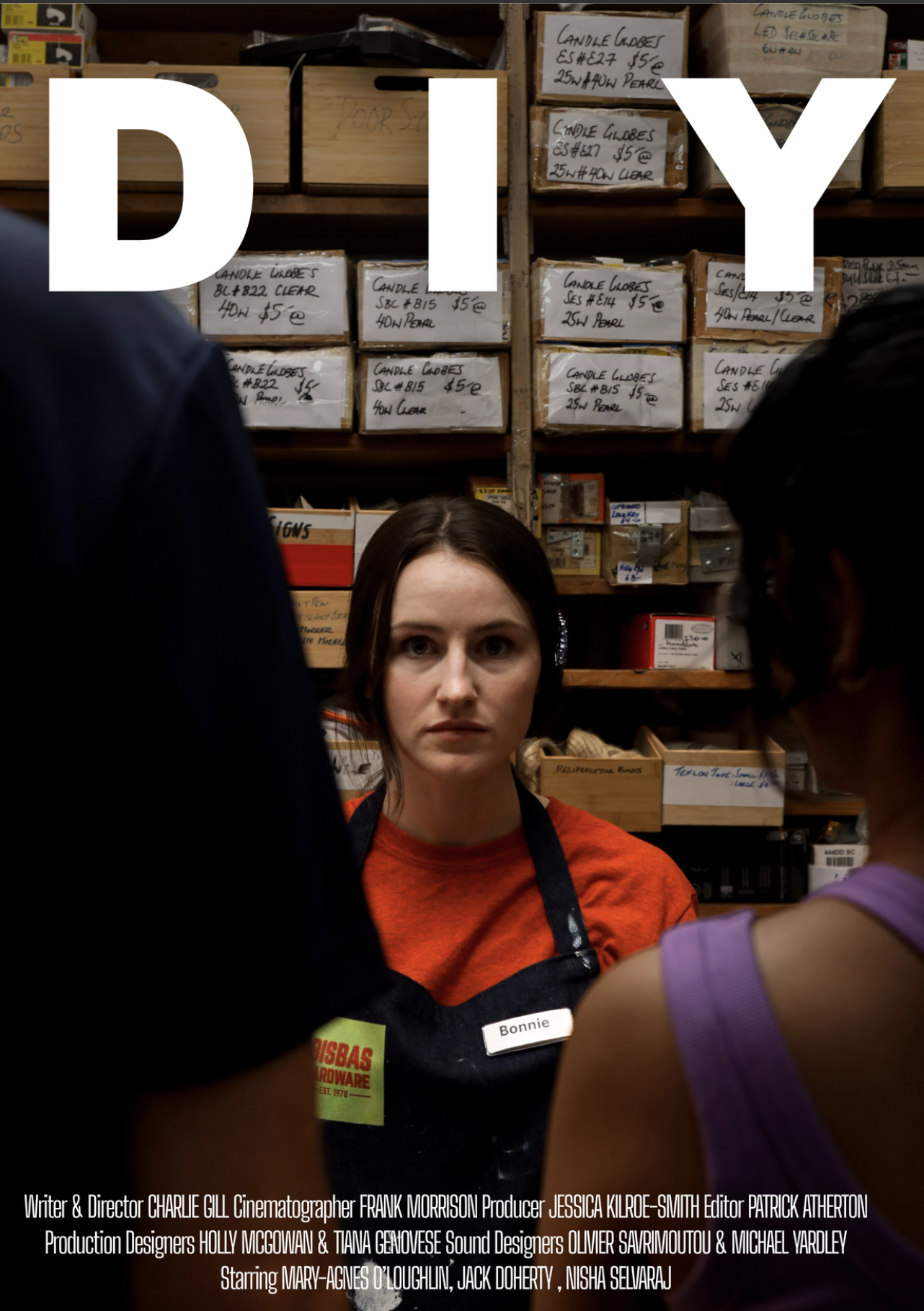 A woman with dark hair, wearing an orange shirt and a black apron with a name tag that says 'Bonnie,' is looking directly at the camera in a workspace with shelves full of boxes labeled with various candle making supplies. Two people are partially visible in the foreground.