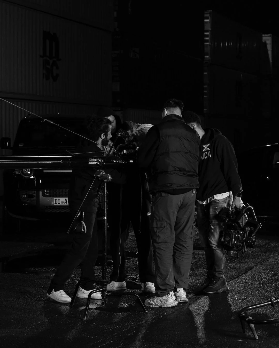 Black and white image of a film crew gathered around a camera in an industrial setting with shipping containers in the background.