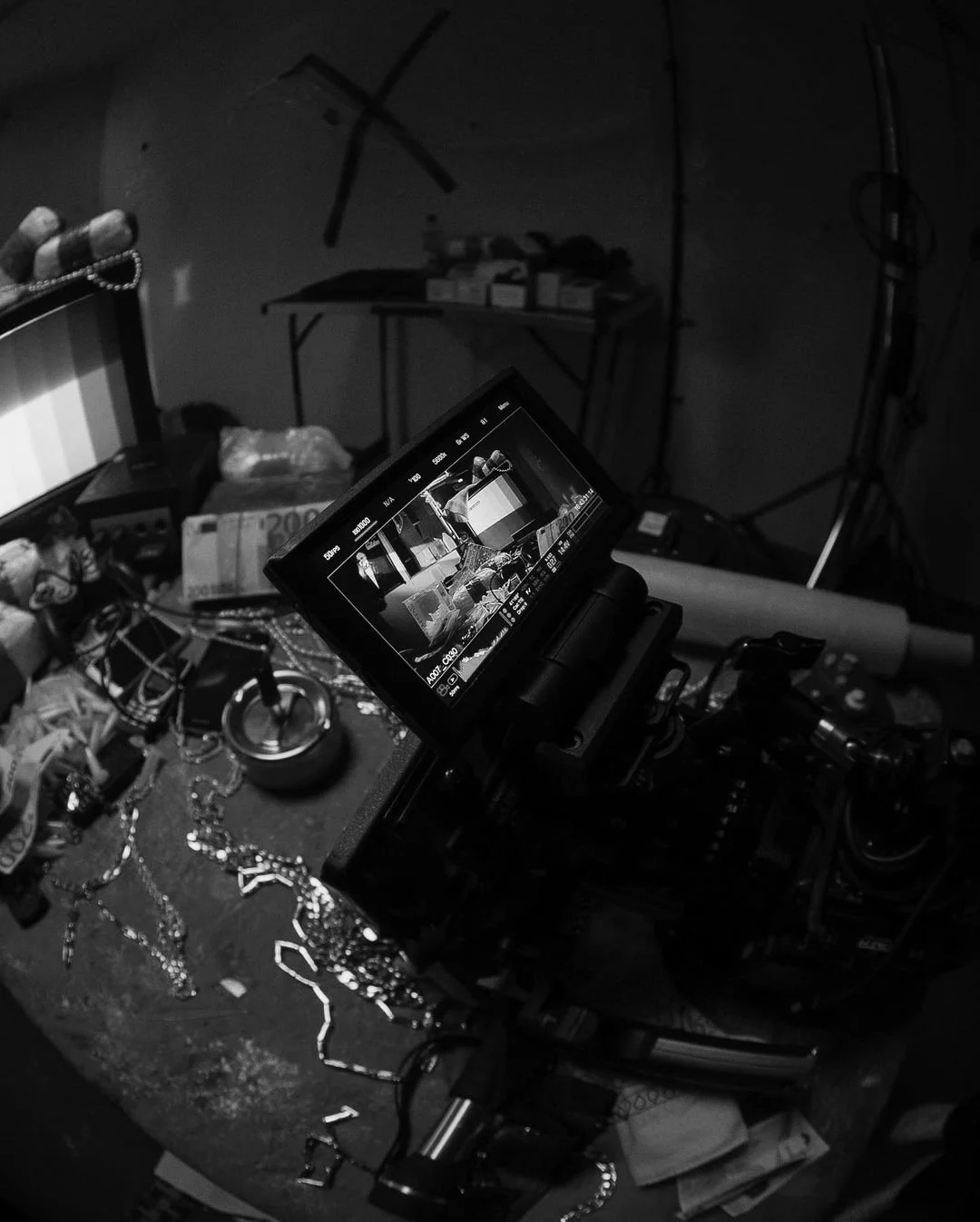 Black and white photo of a cluttered workspace with a video camera in the foreground. The camera's display screen shows a preview of the scene being filmed, featuring various objects and money on a chaotic surface. The background includes a table with boxes and equipment, and wall markings.