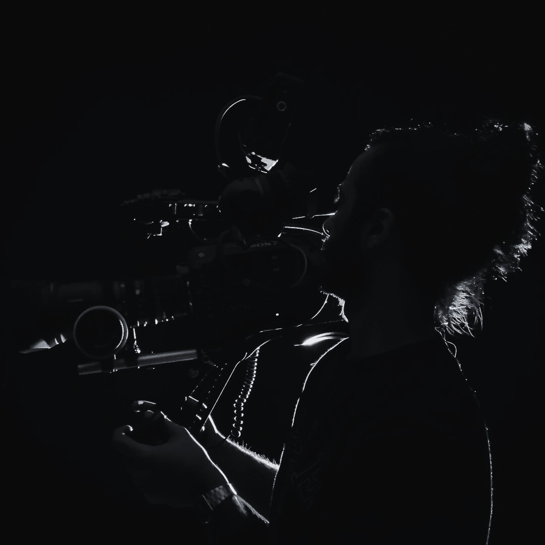 Silhouette of a person operating a professional video camera with a dark background, light outlining their profile and the camera gear.