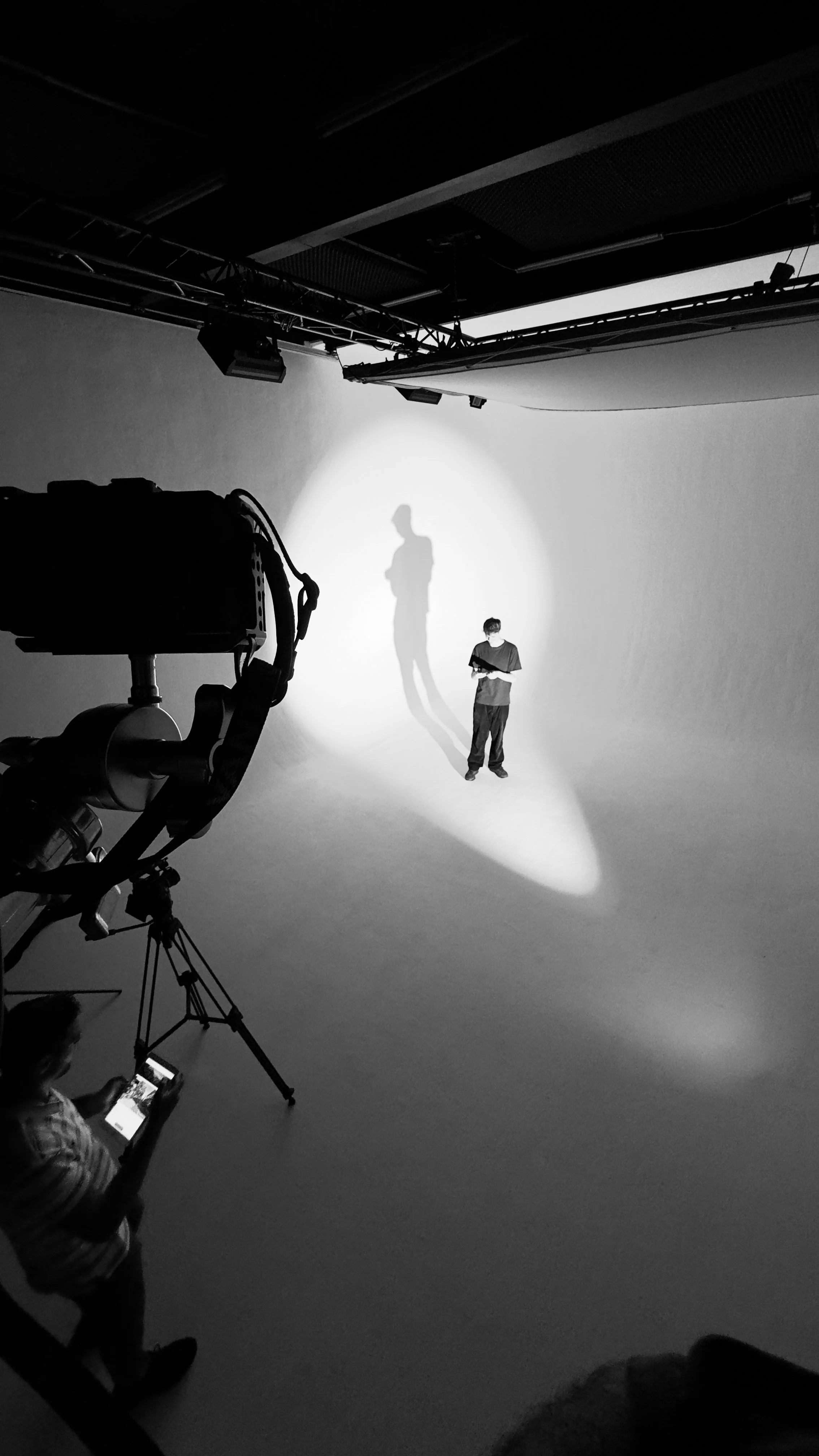 Black and white photo of a man standing in a studio with overhead lighting casting a shadow; a camera on a tripod is in the foreground.