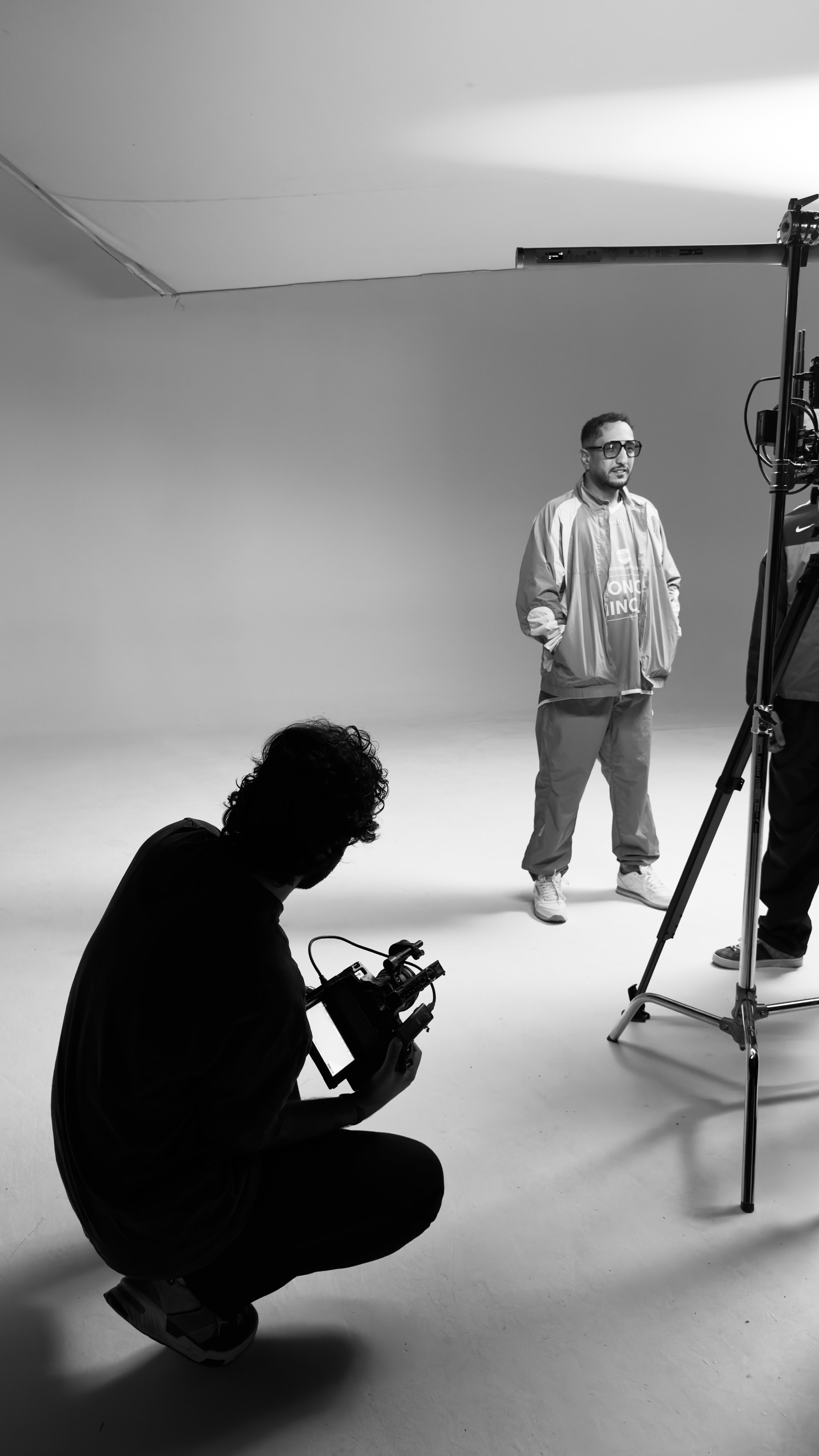 Black and white photo of a film set with a man standing in front of a camera and a camera operator crouching nearby.