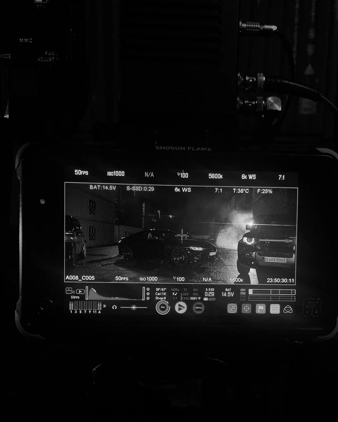 Camera monitor showing a black and white film scene with two parked cars and steam or smoke in the background.