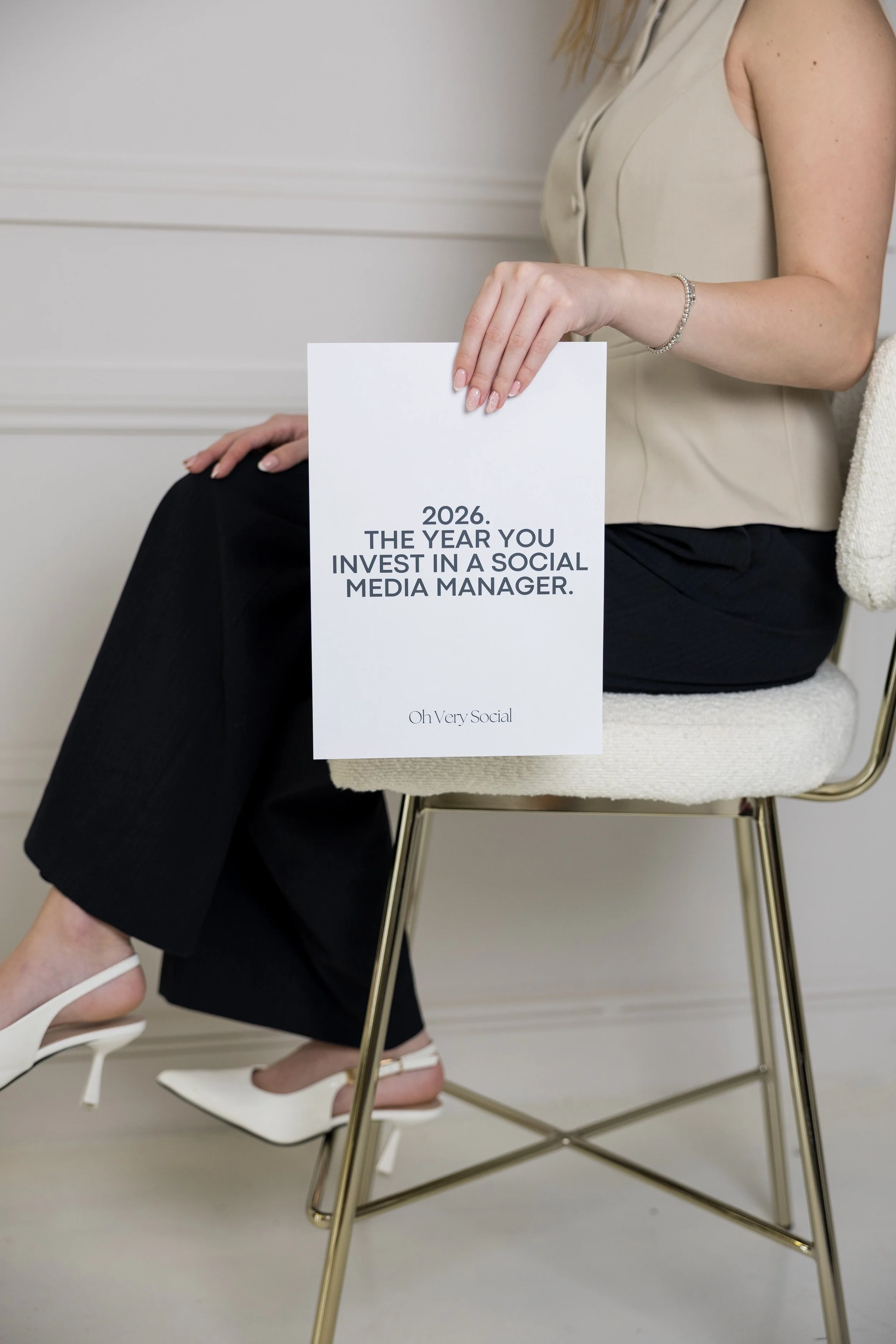 A woman sitting on a white chair with gold legs, holding a sign that says, "2026. The year you invest in a social media manager," with a white wall in the background.