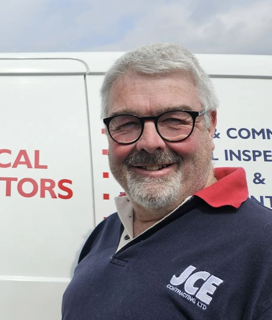 A smiling man with gray hair, glasses, and a beard wearing a navy blue and red collared shirt stands in front of a white vehicle with partially visible text.