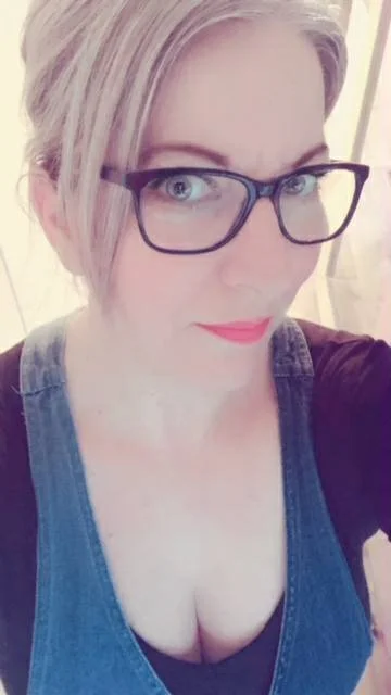 Close-up selfie of a woman with blonde hair, wearing glasses, a dark top with a denim dress, and pink lipstick.