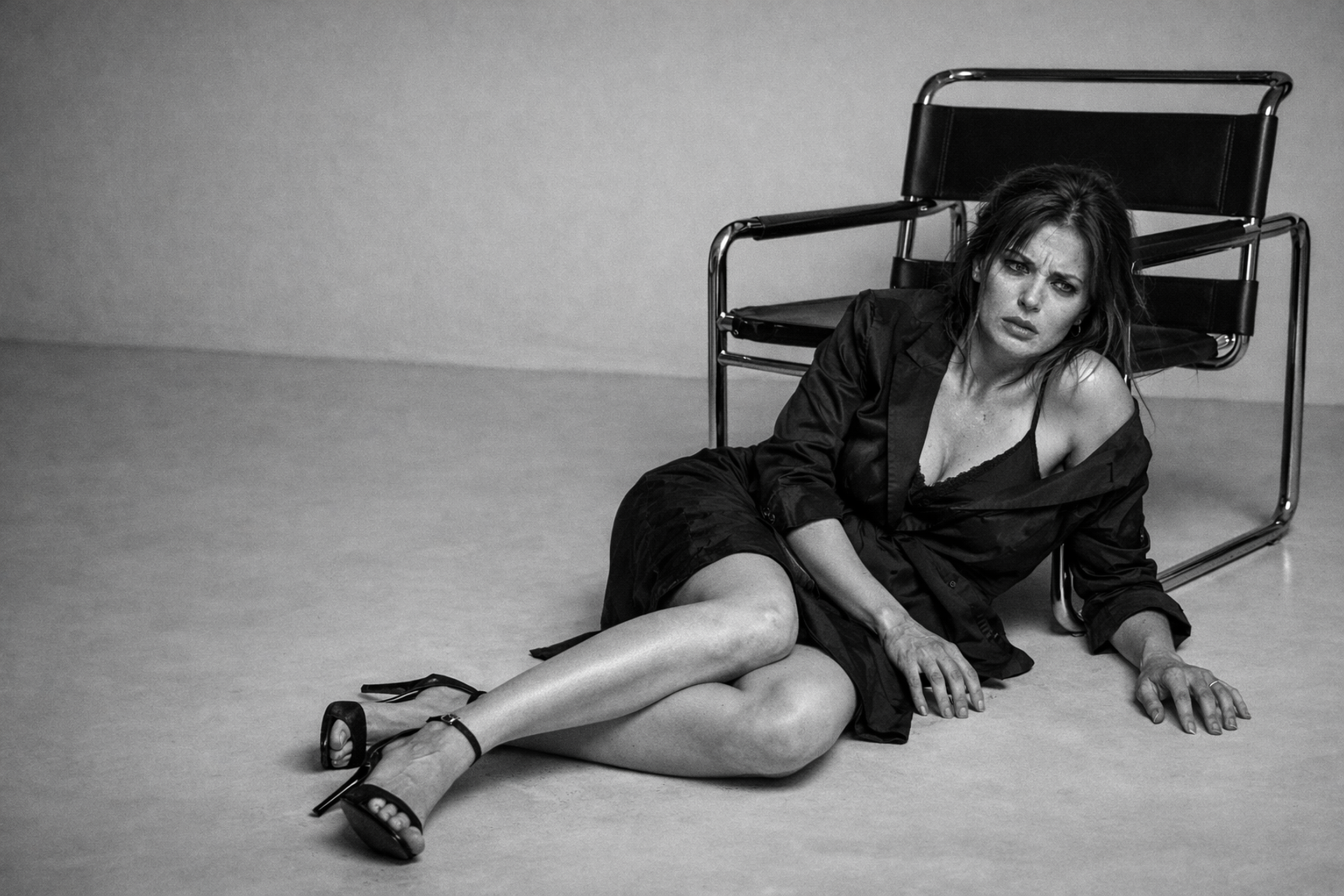 A woman in a black dress and heels sitting on the floor with her back against a chair, looking distressed in a black-and-white photo.