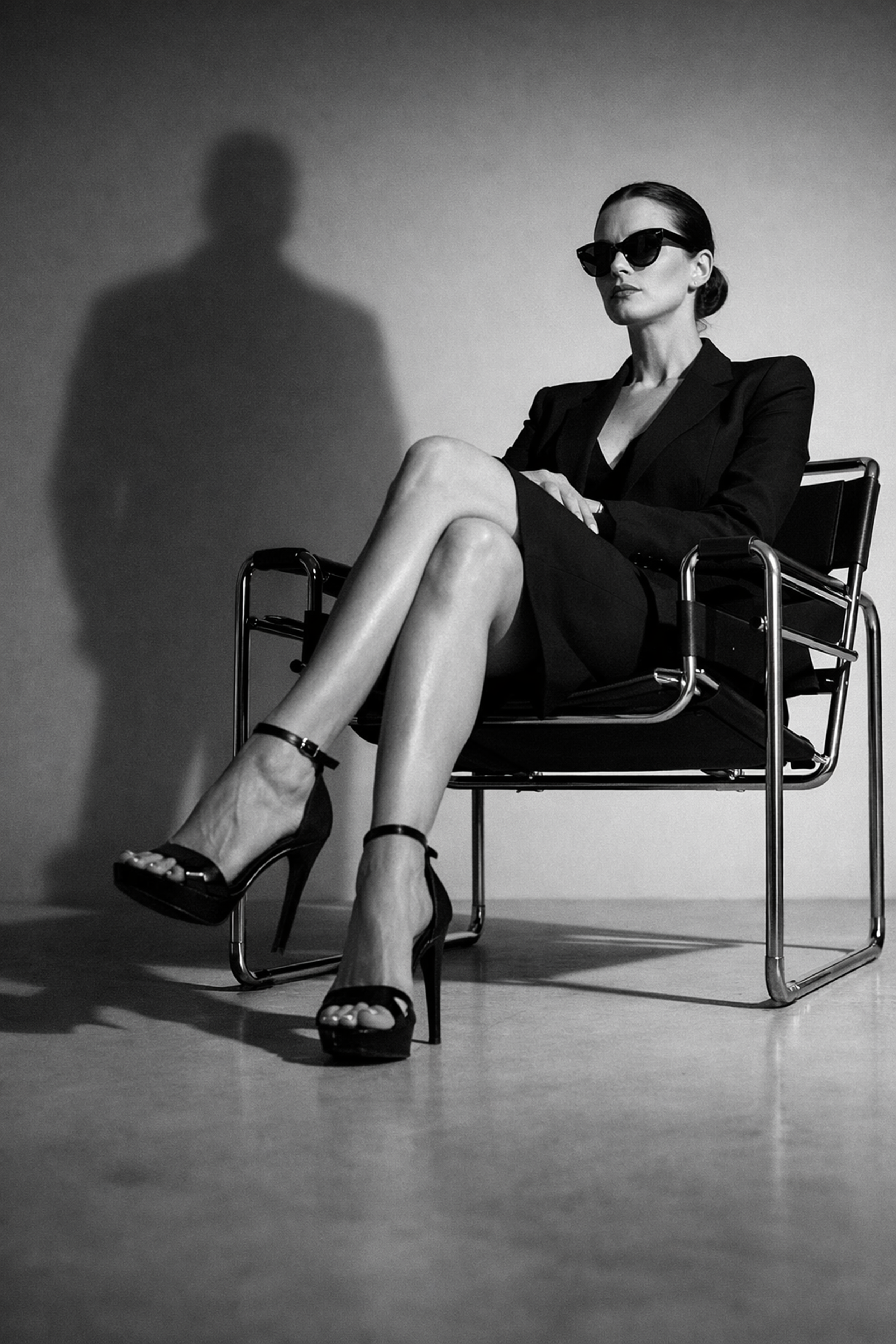 A woman in black attire and sunglasses, sitting with her legs crossed in a chair, casting a shadow on the wall behind her.