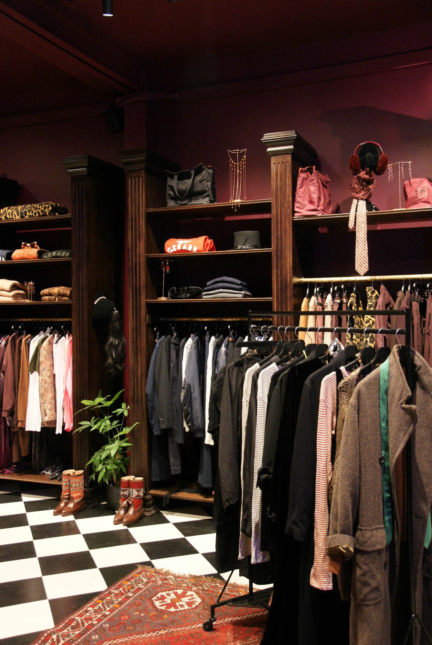 Gerard Antwerp clothing store with racks of jackets, shirts, and dresses, shelves with bags and accessories, and a potted plant on a checkered floor area rug.