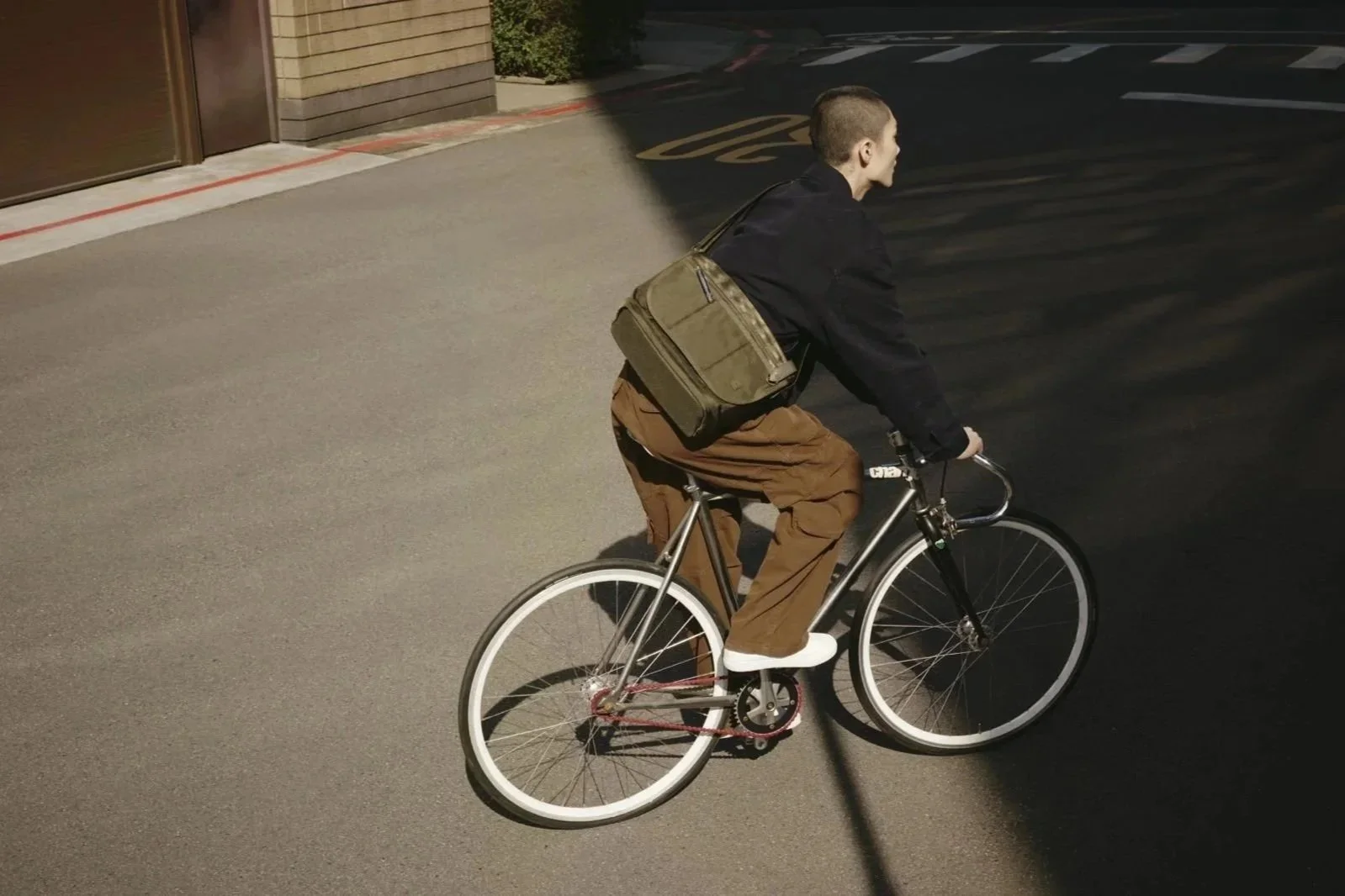 A young man with a backpack riding a bicycle on a city street during daytime, with a shadow cast over part of the road and sidewalk.