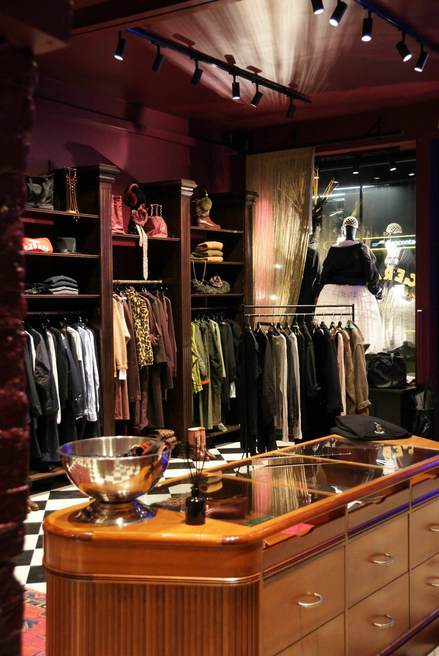 Interior of a Gerard Antwerp clothing store with racks of clothes, shelves with accessories, and a display window. The store has a warm, vintage decor with wooden furniture and checkered flooring.