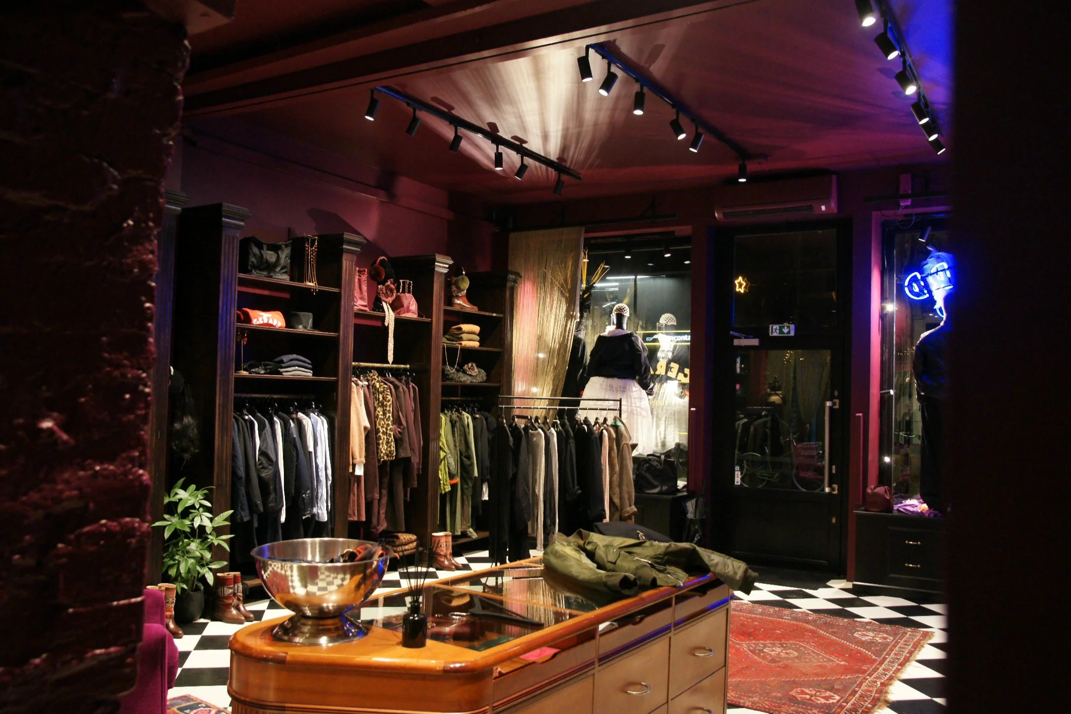Interior of a retail clothing store with warm lighting, wood cabinets, clothing racks, and mannequins dressed in fashion. There are decorative elements, neon signs, and a checkered floor.