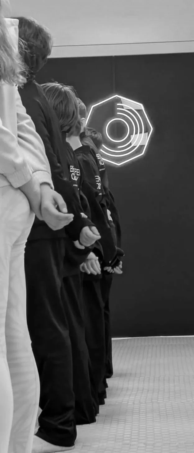 Mehrere Menschen stehen nebeneinander in einer Reihe, alle mit den Händen auf dem Rücken, in einem Raum mit einer dunklen Wand und einer Neon-Hexagon-Design an der Wand im Hintergrund.