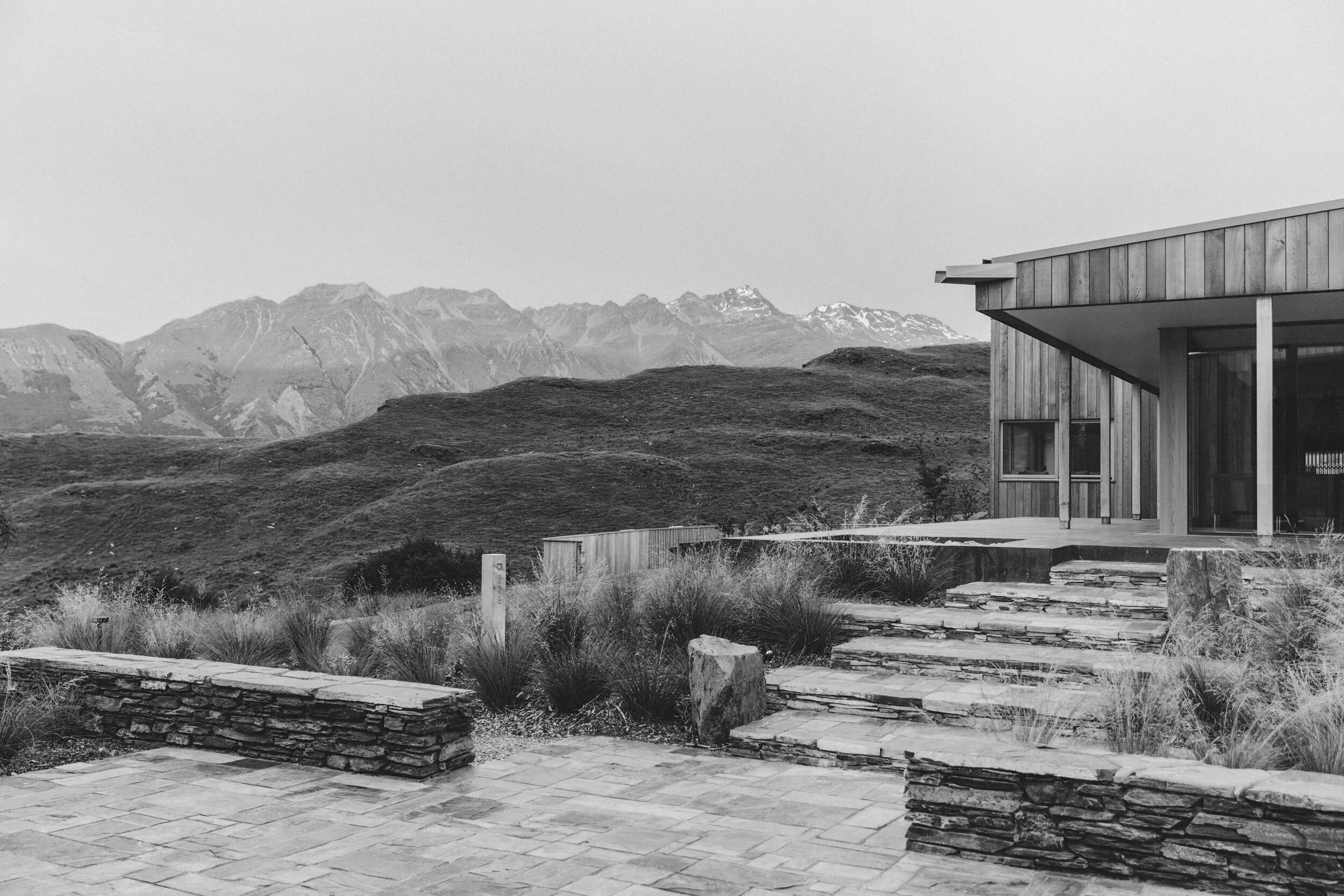 Ein modernes Holzhaus mit steinernen Terrassen und Treppen, umgeben von Bergen im Hintergrund, in Schwarz-Weiß.