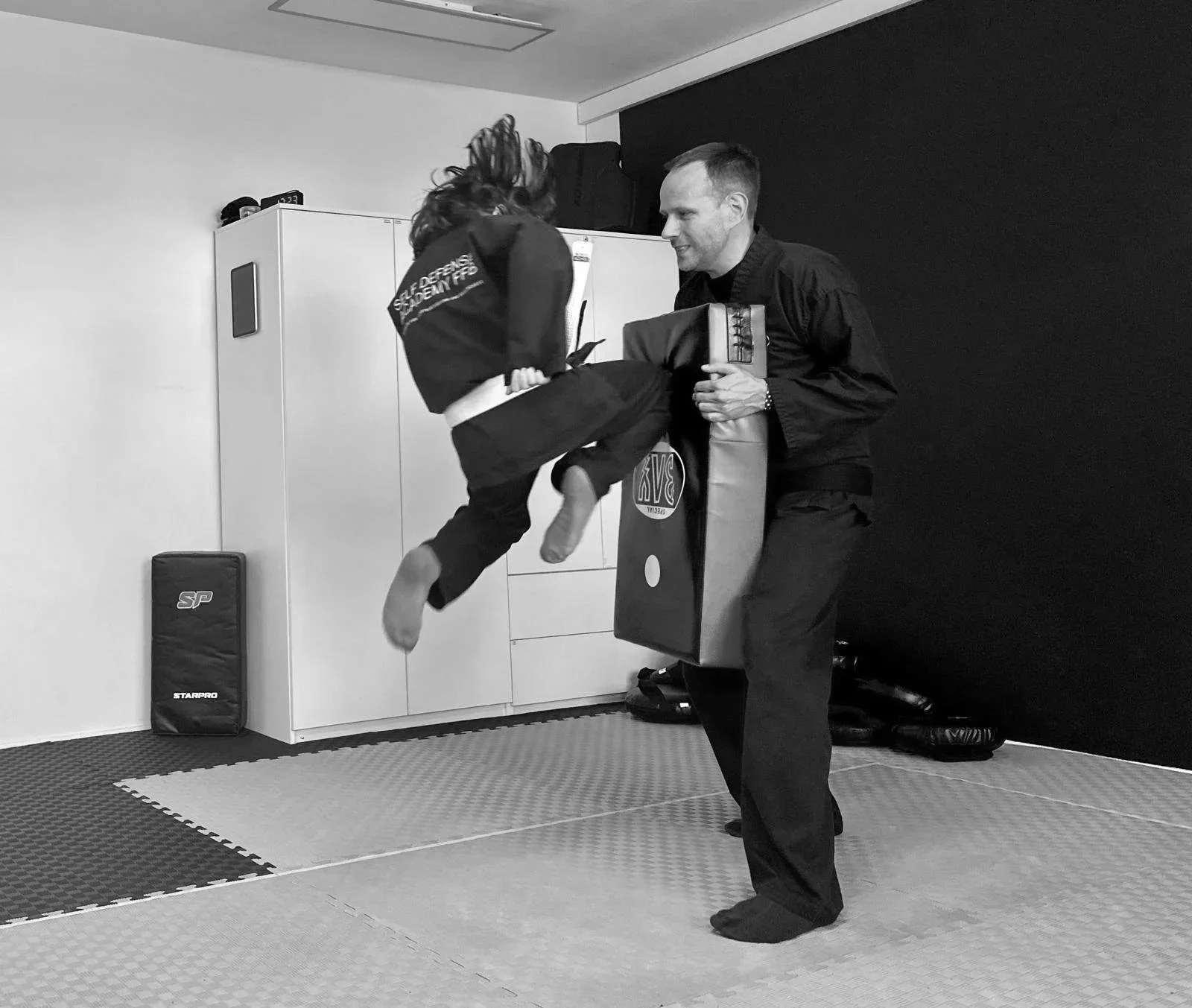 Ein Mann trainiert mit einem Kind in einer Kampfsportschule. Das Kind trägt einen Gi und springt in der Luft, während der Mann es mit Schutzausrüstung abwehrt.