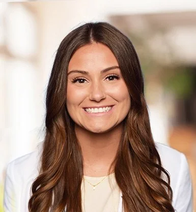 A woman with long brown hair smiling, wearing a white blazer and a light-colored top, with a blurred outdoor background.