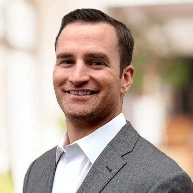 A smiling man in a gray suit and white shirt, outdoors.
