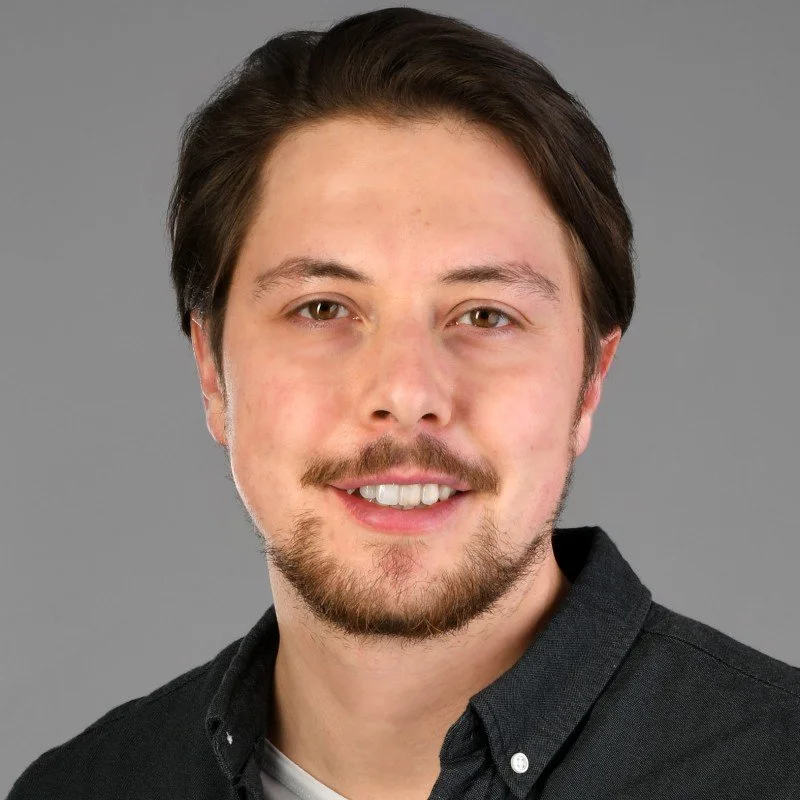 Porträt eines jungen Mannes (Dr. med. Timo Trampert) mit dunklen Haaren, Bart und einem schwarzen Hemd vor grauem Hintergrund.