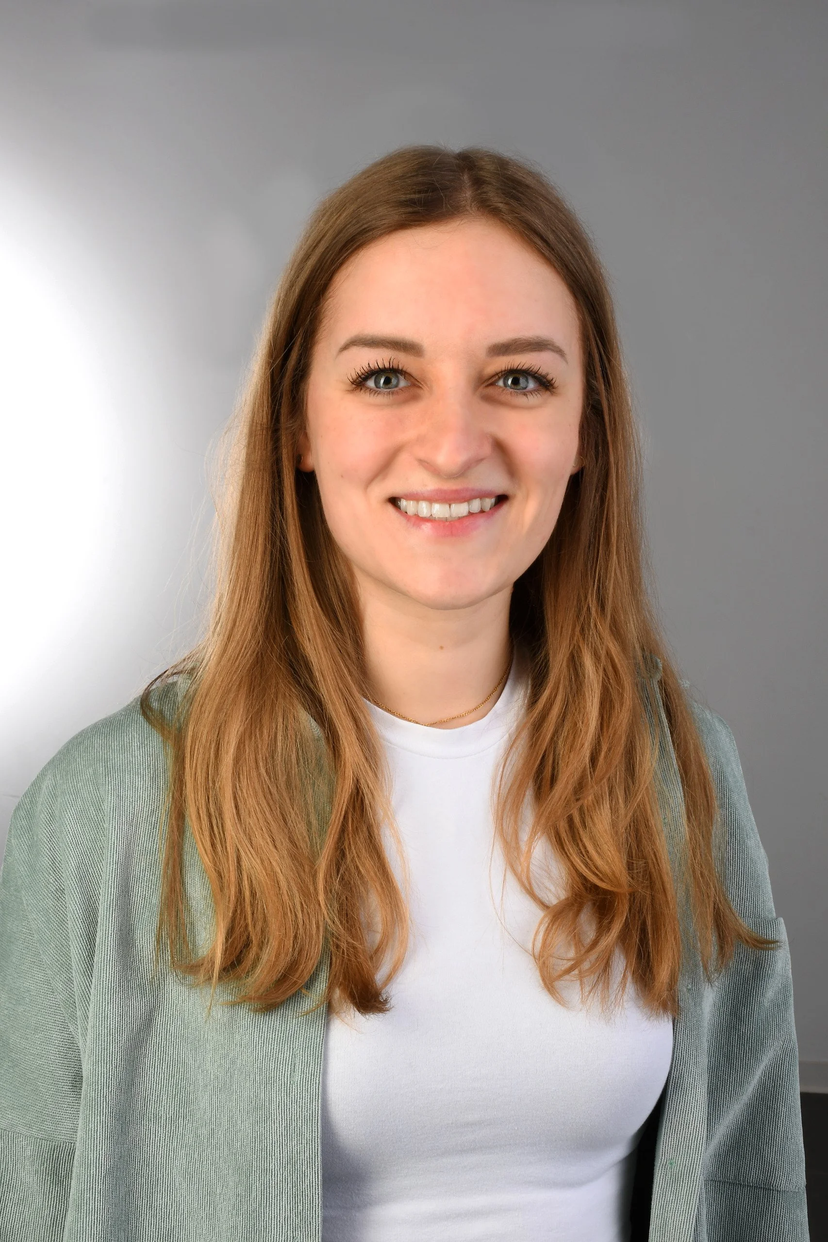 Junge Frau (Tamira Trampert) mit langen braunen Haaren, blauen Augen, trägt weißen T-Shirt und grünen Pullover, lächelnd vor grauem Hintergrund.
