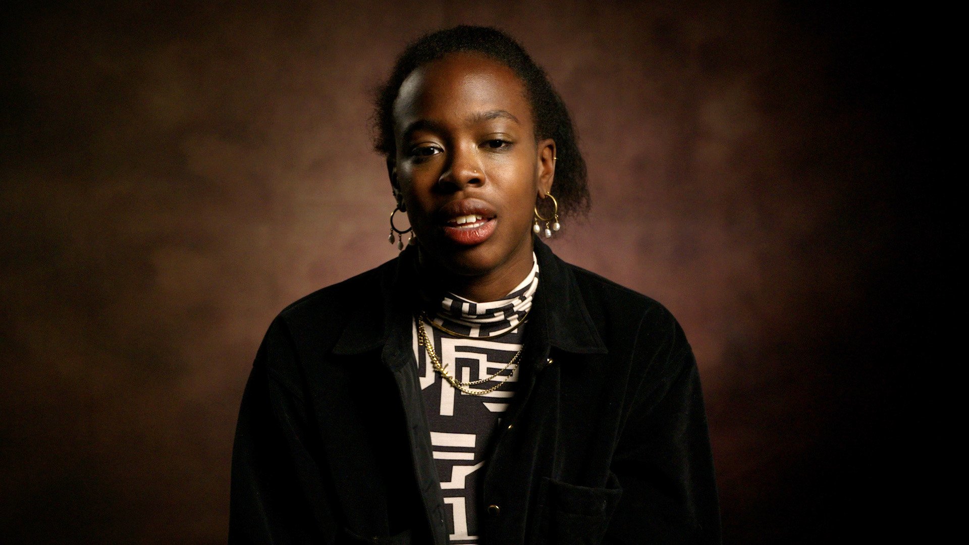 Portrait of a young woman with dark skin, short curly hair, wearing hoop earrings, a patterned turtleneck, and a black jacket, against a dark brown background.