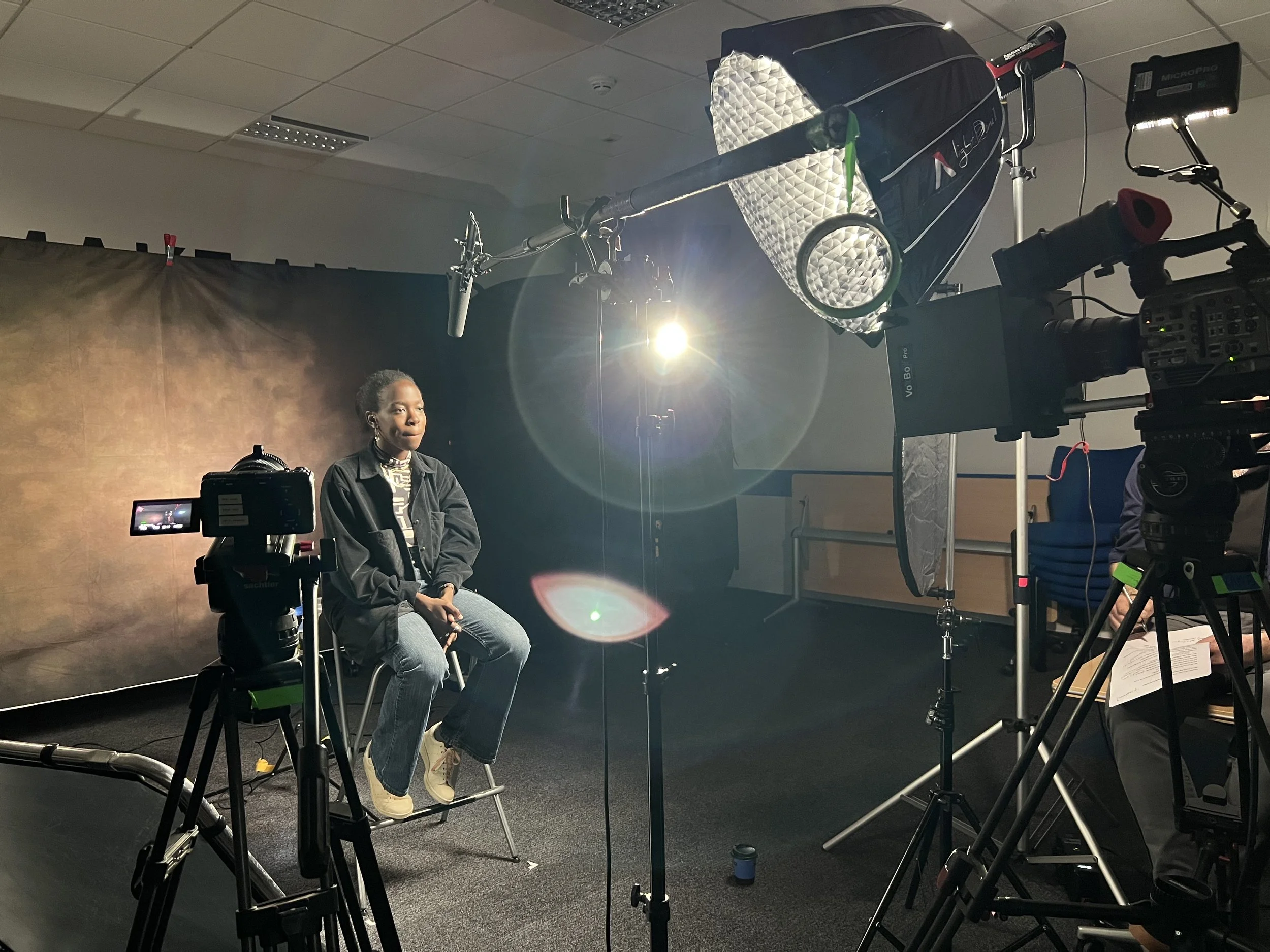 A woman sitting on a stool during a video shoot or interview, surrounded by professional filming equipment in a studio with a dark backdrop.