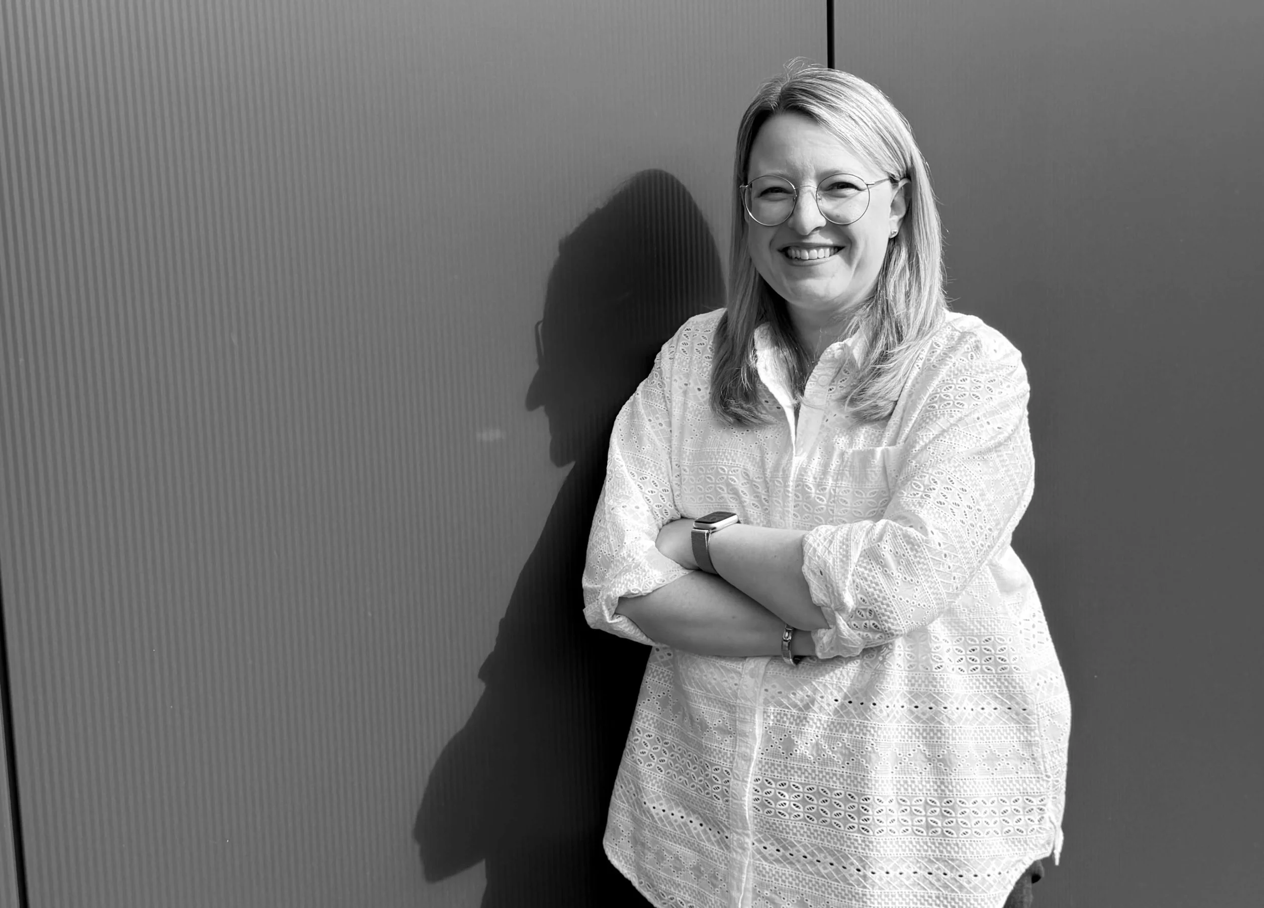 Schwarz-Weiß-Foto einer lachenden Frau mit Brille, die vor einer Wand steht und ihre Arme verschränkt hat.