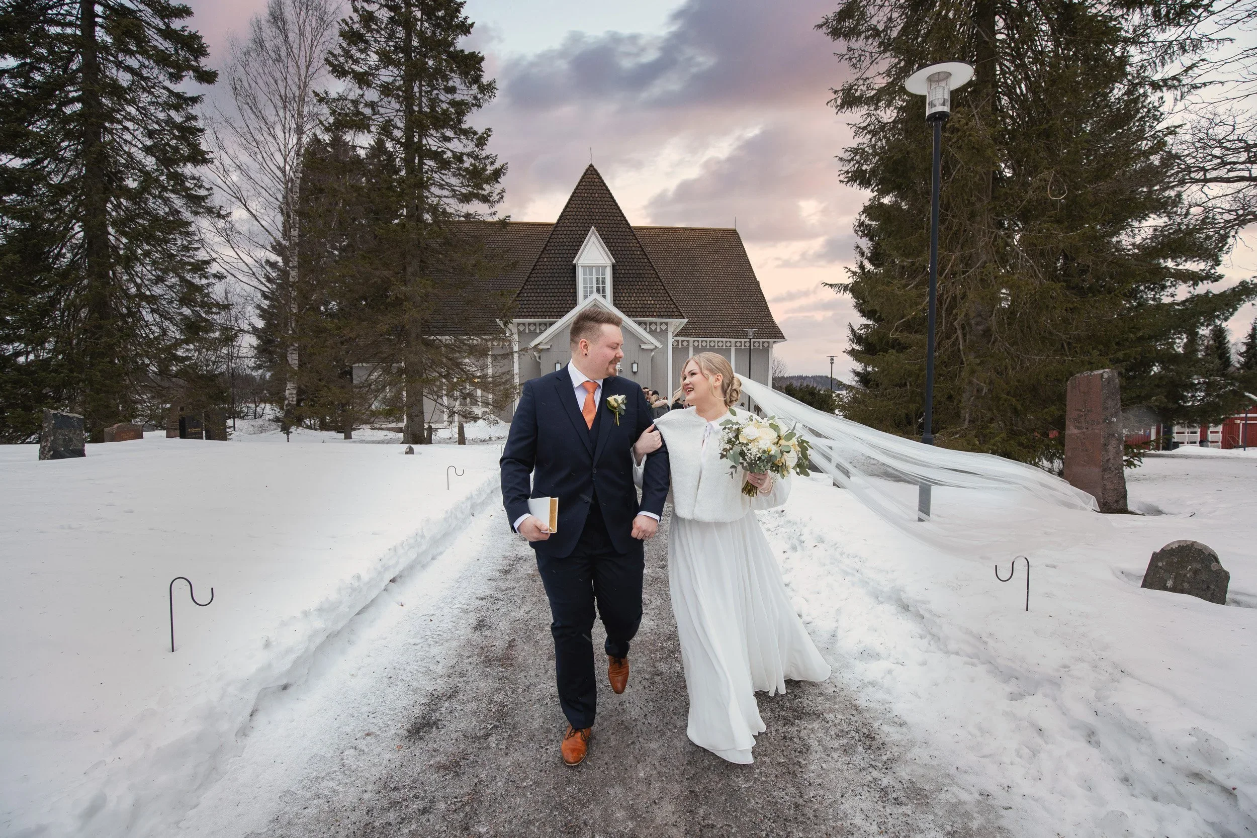 Hääpari kävelee lumisella tiellä kirkon edessä, nainen pitää kukkakimppua.