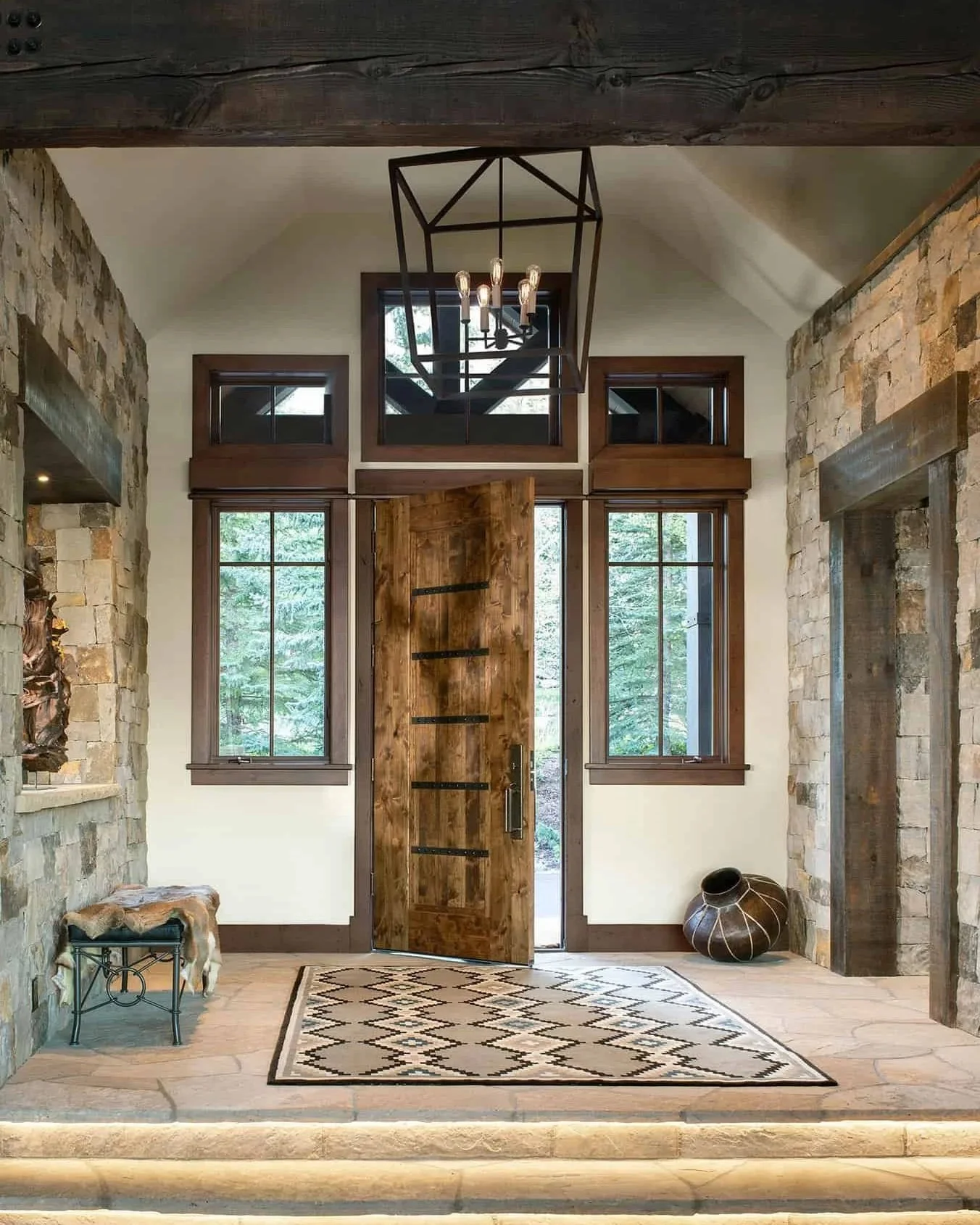 Rustic interior entryway with a honey-colored wooden front door, large windows, stone walls, a patterned rug, decorative vase, and a geometric chandelier.