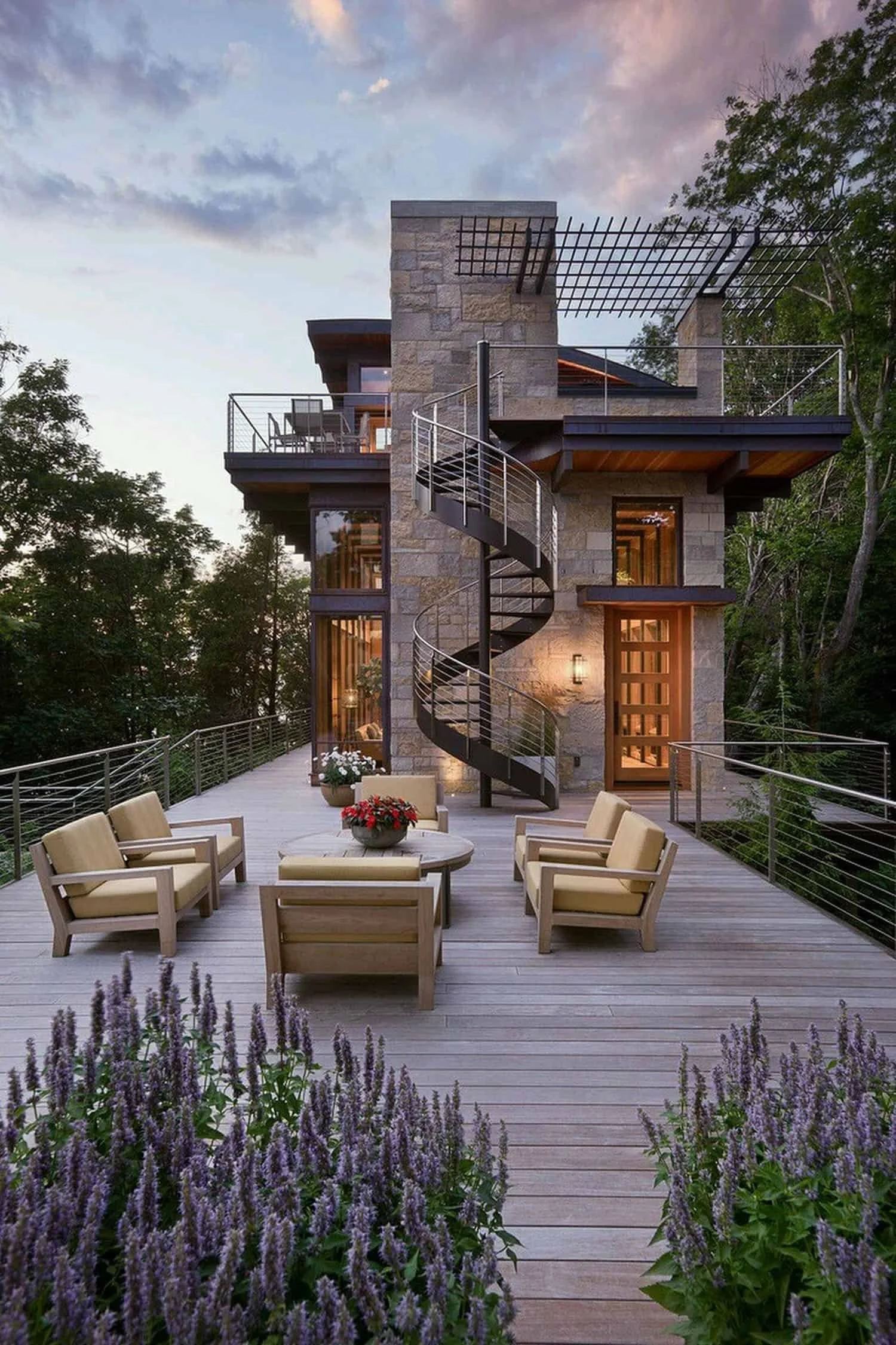 Colorado mountain second home with a wooden deck, outdoor seating arrangements, colorful flowers, with a spiral staircase and lush green trees in the background at sunset