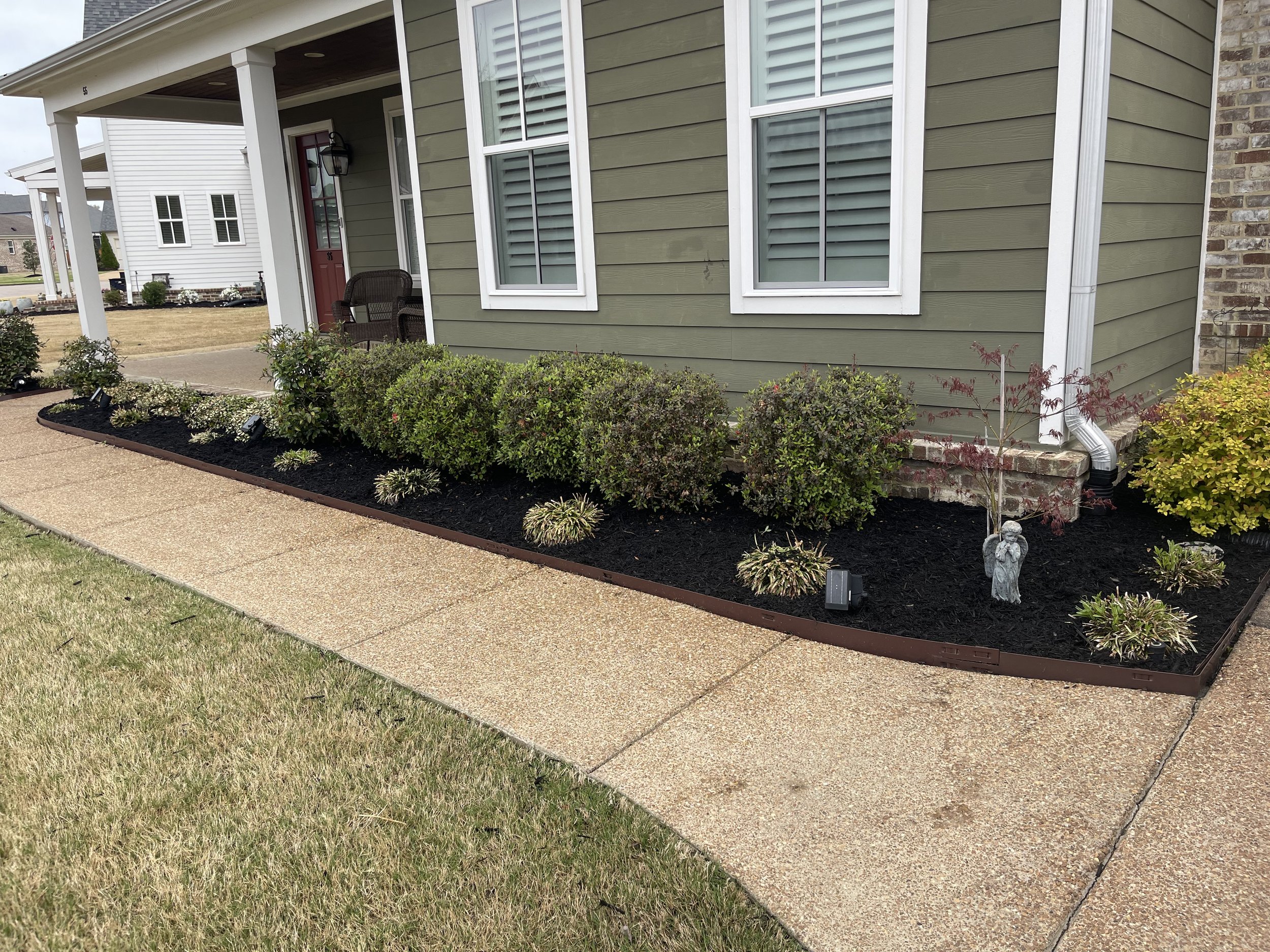Front yard garden with neatly trimmed bushes, small decorative plants, and a statue of an angel, bordered by a brown metal edging, in Piperton, Tennessee.