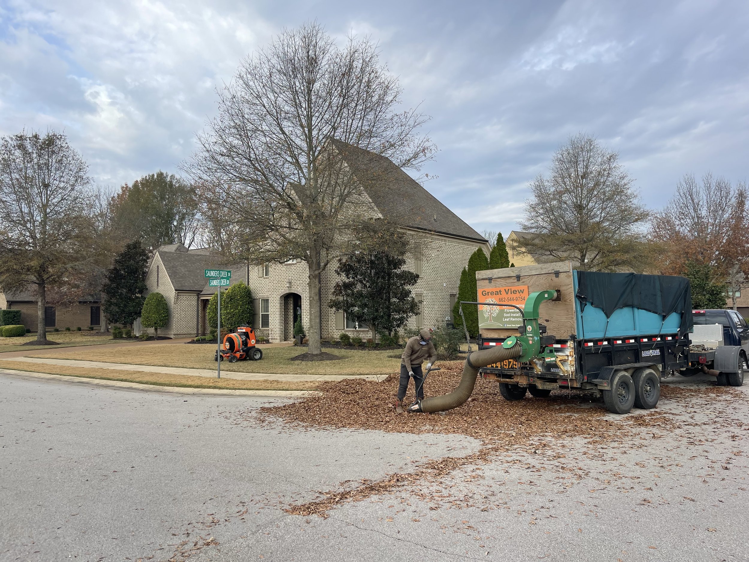 Worker using a leaf blower to clear fallen leaves from the street in front of a residential house during autumn.