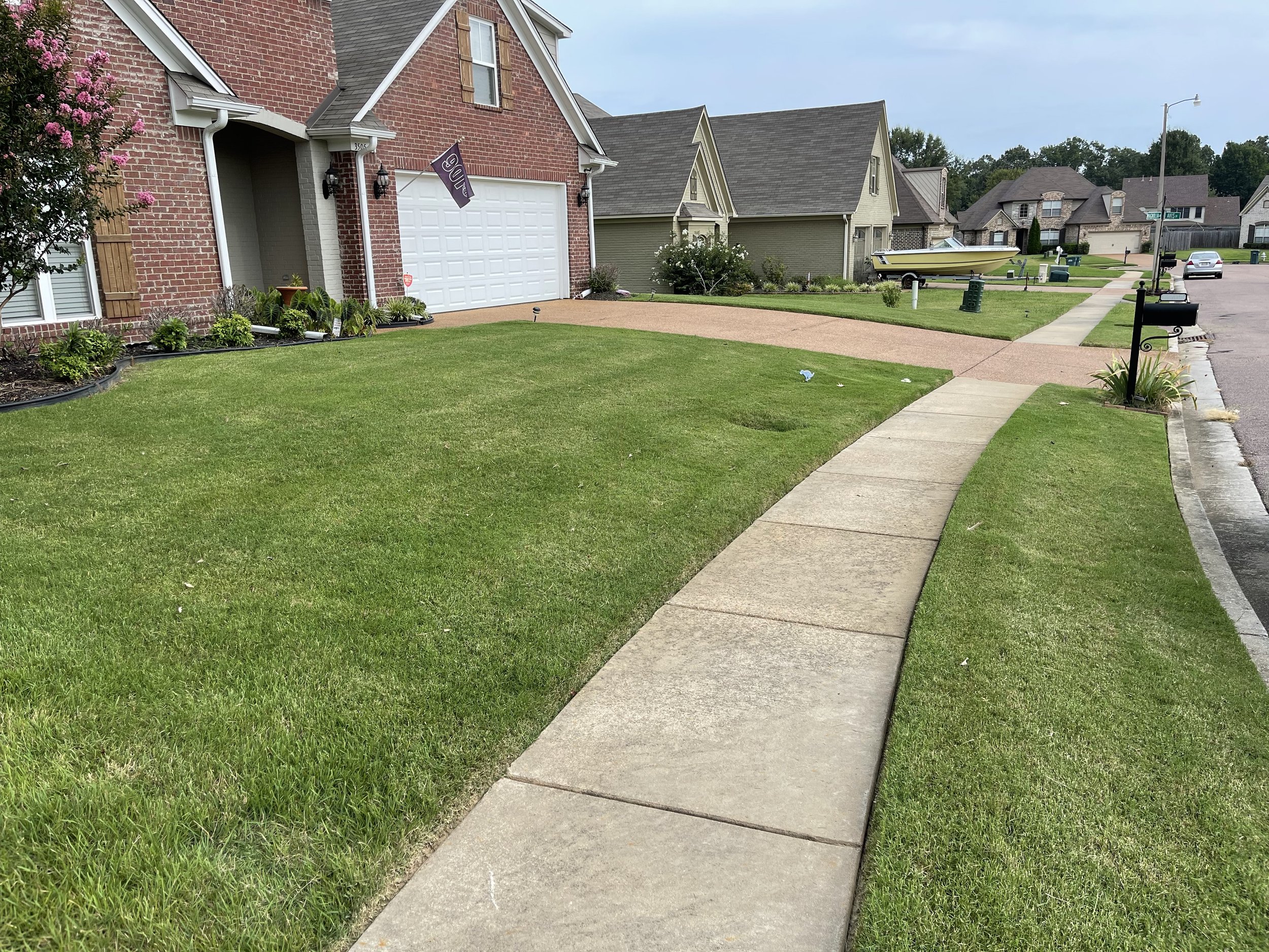 View of a suburban neighborhood sidewalk and front yards with green grass, houses, and boats, on a cloudy day.