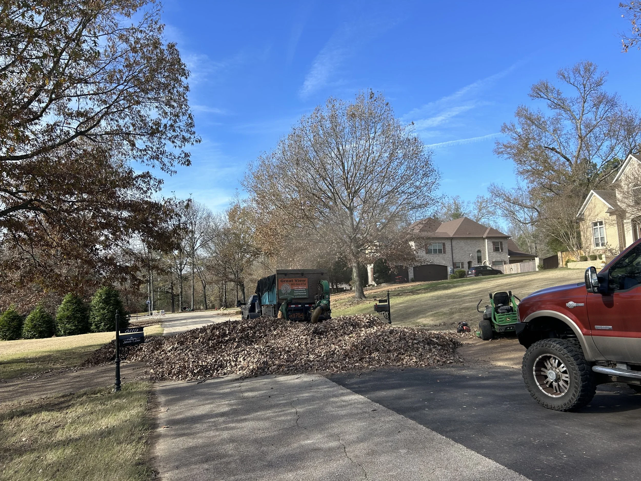 Yard work scene with leaves piled in front of a house on a street. A green leaf blower and a truck with a sign are nearby, with trees and houses in the background under a blue sky.