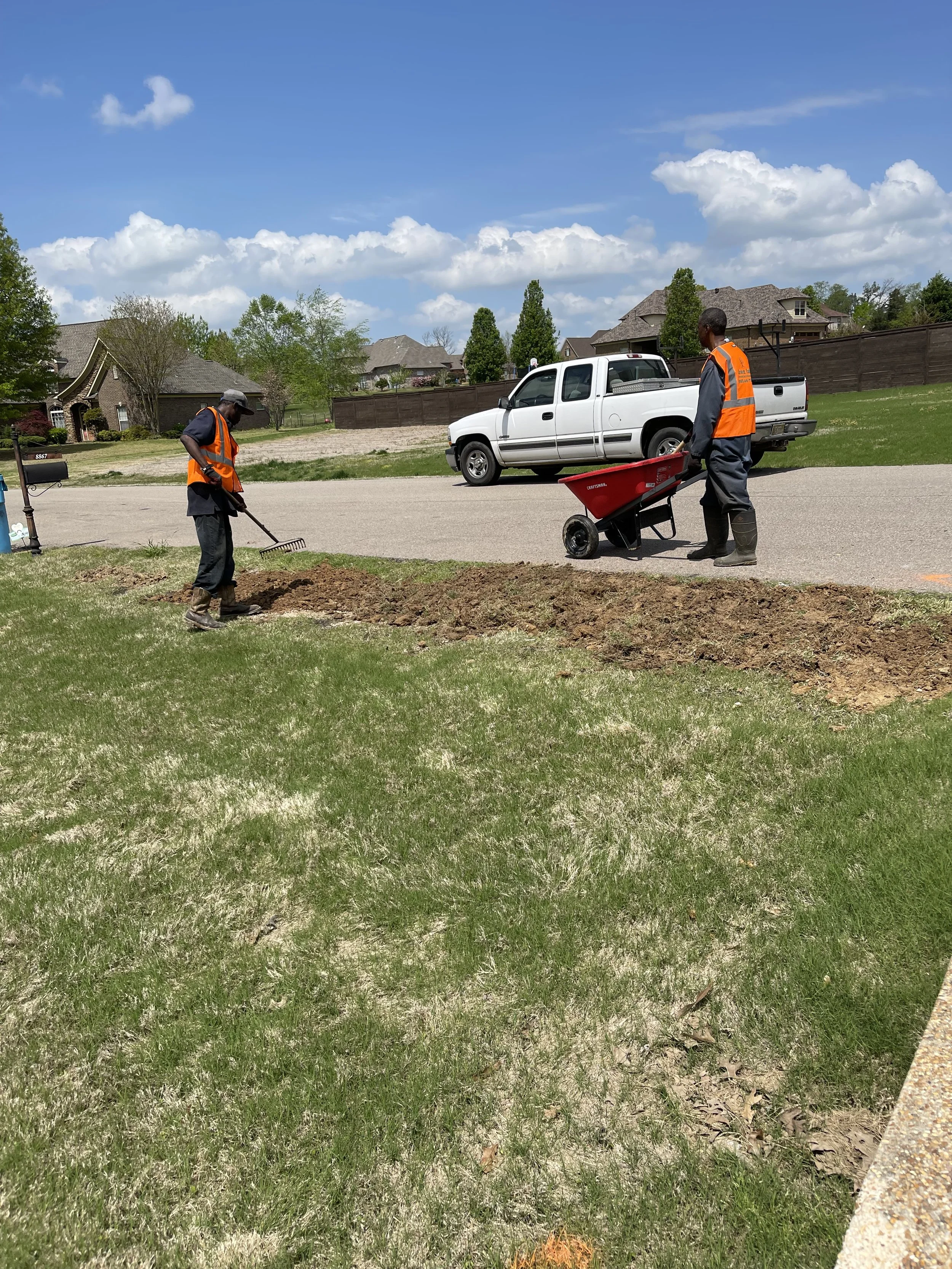 Two workers in orange safety vests and dark clothing yard work on a grassy lawn in a suburban neighborhood, one with a rake and the other with a wheelbarrow near a street, with houses and trees in the background under a partly cloudy sky.