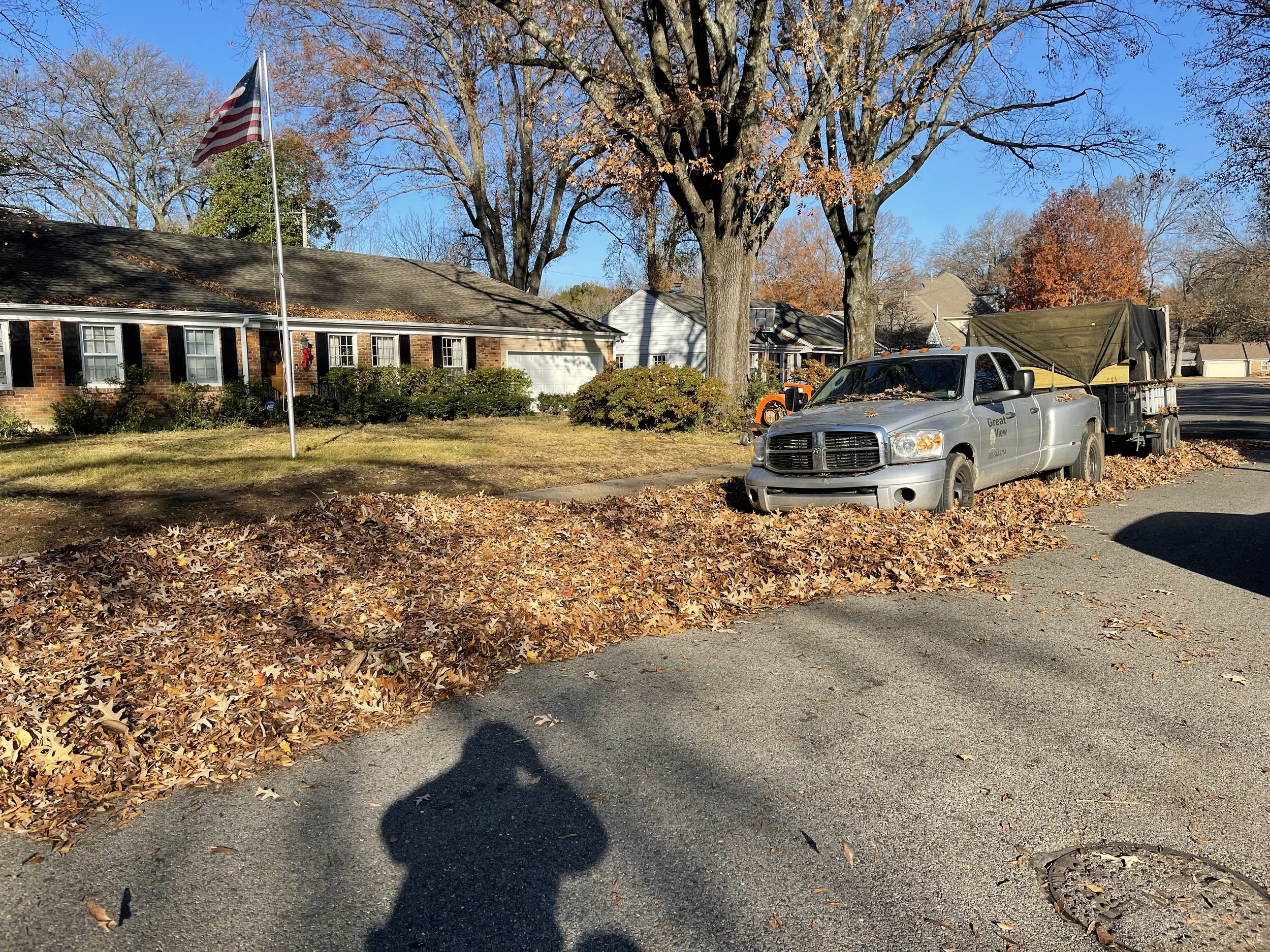 Residential street scene with a house, a flagpole flying the American flag, a large tree without leaves, a gray pickup truck parked on the curb, and a pile of fallen autumn leaves on the road and grass.