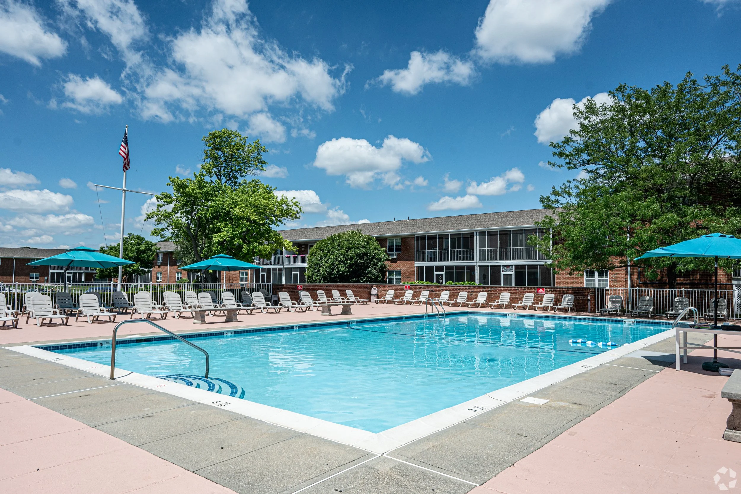 An outdoor swimming pool area with lounge chairs, blue umbrellas, and a residential brick building in the background under a partly cloudy sky.