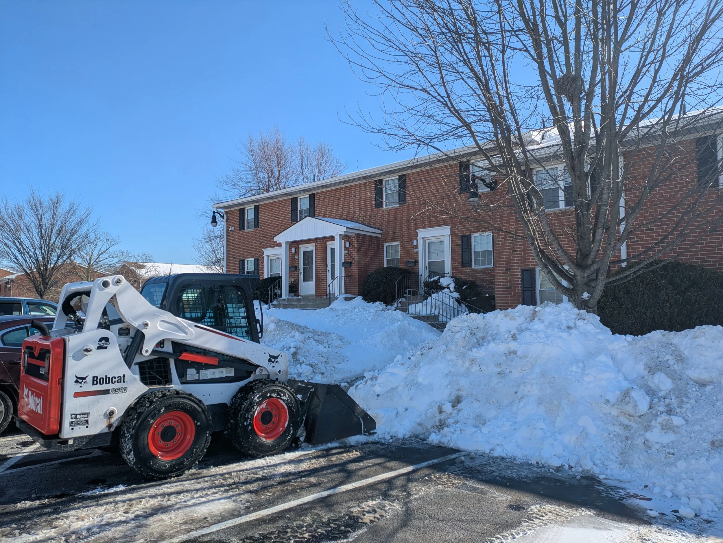 Bobcat snowplow in a plowed parking lot in front of red brick apartment building with piled up snow.