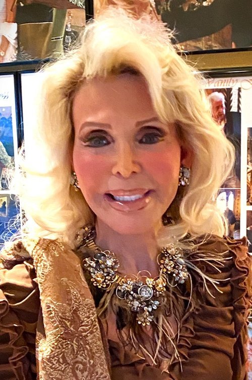 A woman with blonde hair styled in voluminous waves, wearing large earrings, a statement necklace, and a brown, ornate dress, smiling at the camera.