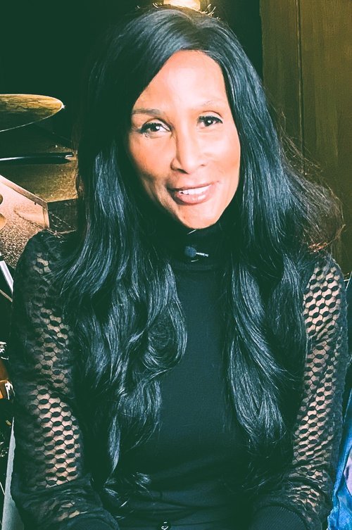 Woman with long black hair wearing a black top with lace sleeves, smiling indoors.