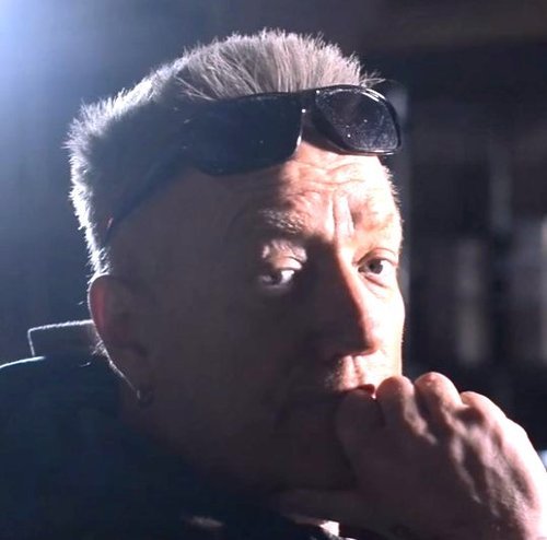 Close-up of a man with sunglasses on his head, resting his chin on his hand, in a dimly lit environment.