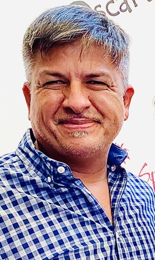 Man with short, graying hair, wearing a blue and white checkered shirt, smiling in front of a white background with partial text.