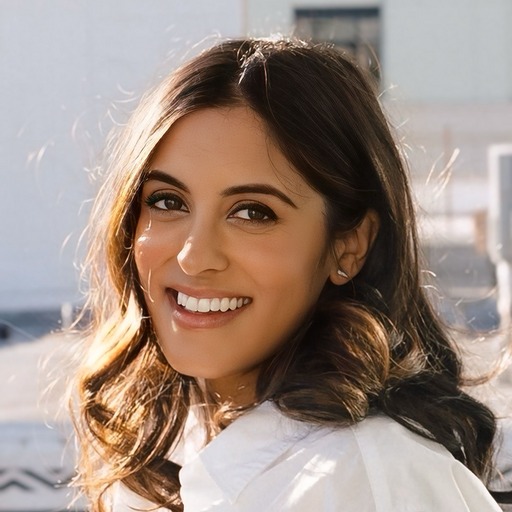 Young woman with long, wavy brown hair, smiling outdoors with sunlight on her face, wearing a white shirt.