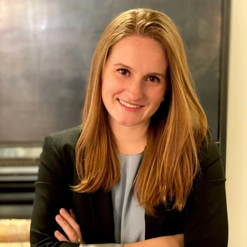 A young woman with long, straight red hair, smiling and crossing her arms, wearing a light grey top and a black blazer, in an indoor setting with neutral background.