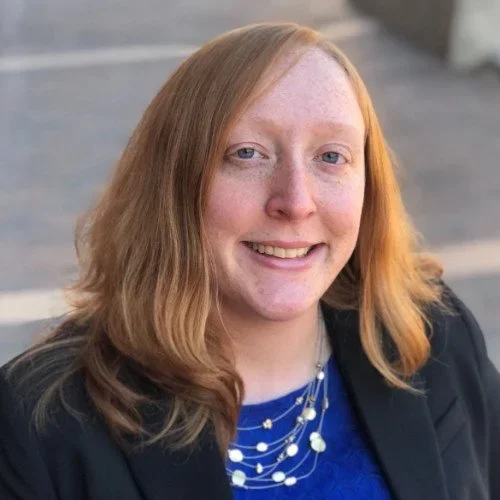 A woman with red hair and freckles smiling outdoors.