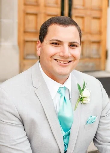 Smiling man in a light gray suit with a light blue tie and boutonniere, outdoors with wooden background.