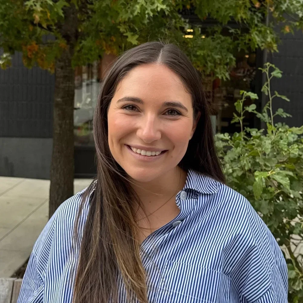 A woman with long brown hair and a striped blue and white shirt standing outdoors, smiling with trees and plants in the background.