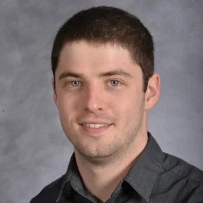 Portrait of a young man with short dark hair, light skin, wearing a dark gray button-up shirt, smiling against a gray background.
